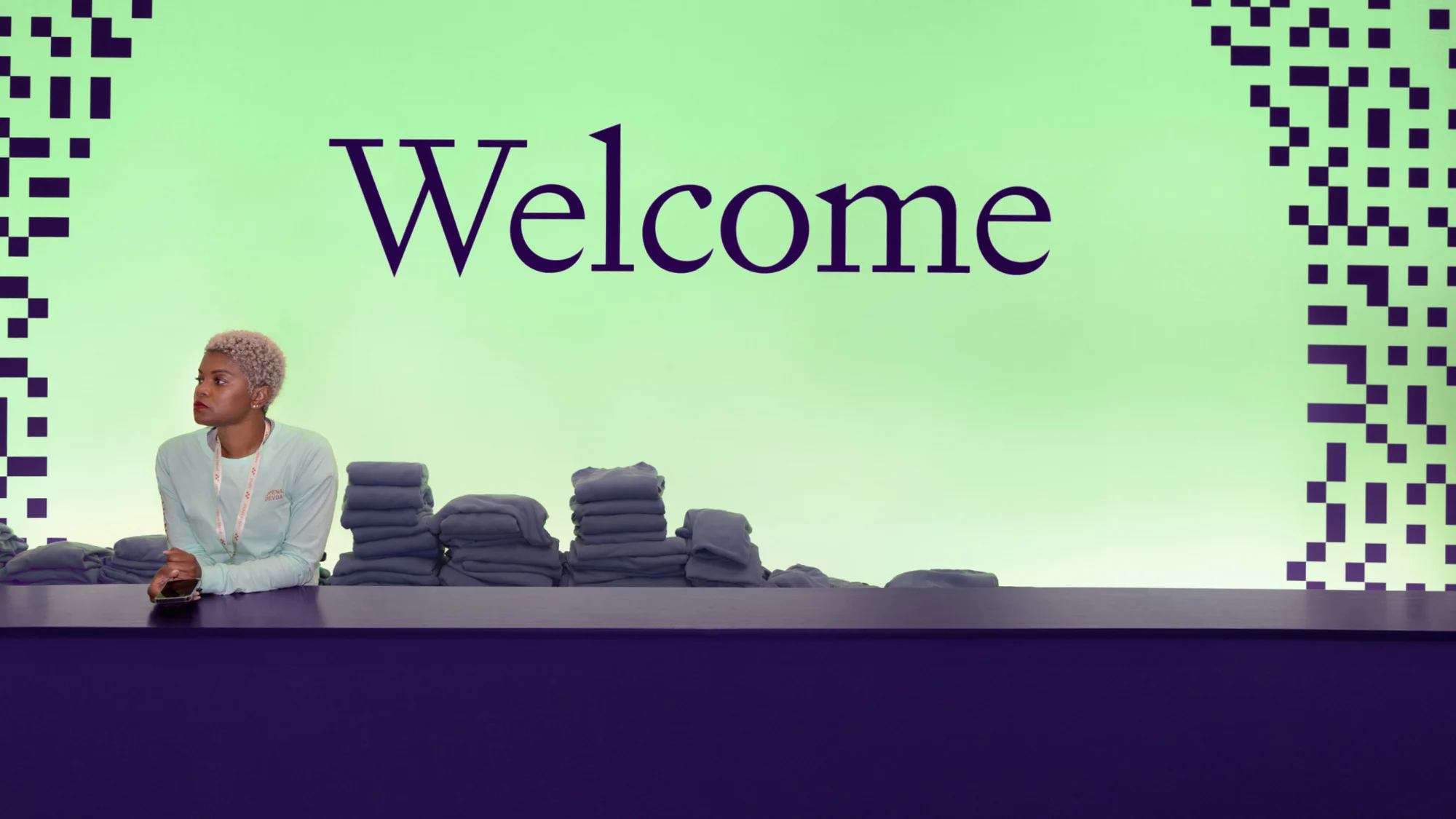 Adult female standing behind a front desk with towels against a graphic green background with the word Welcome in purple font.