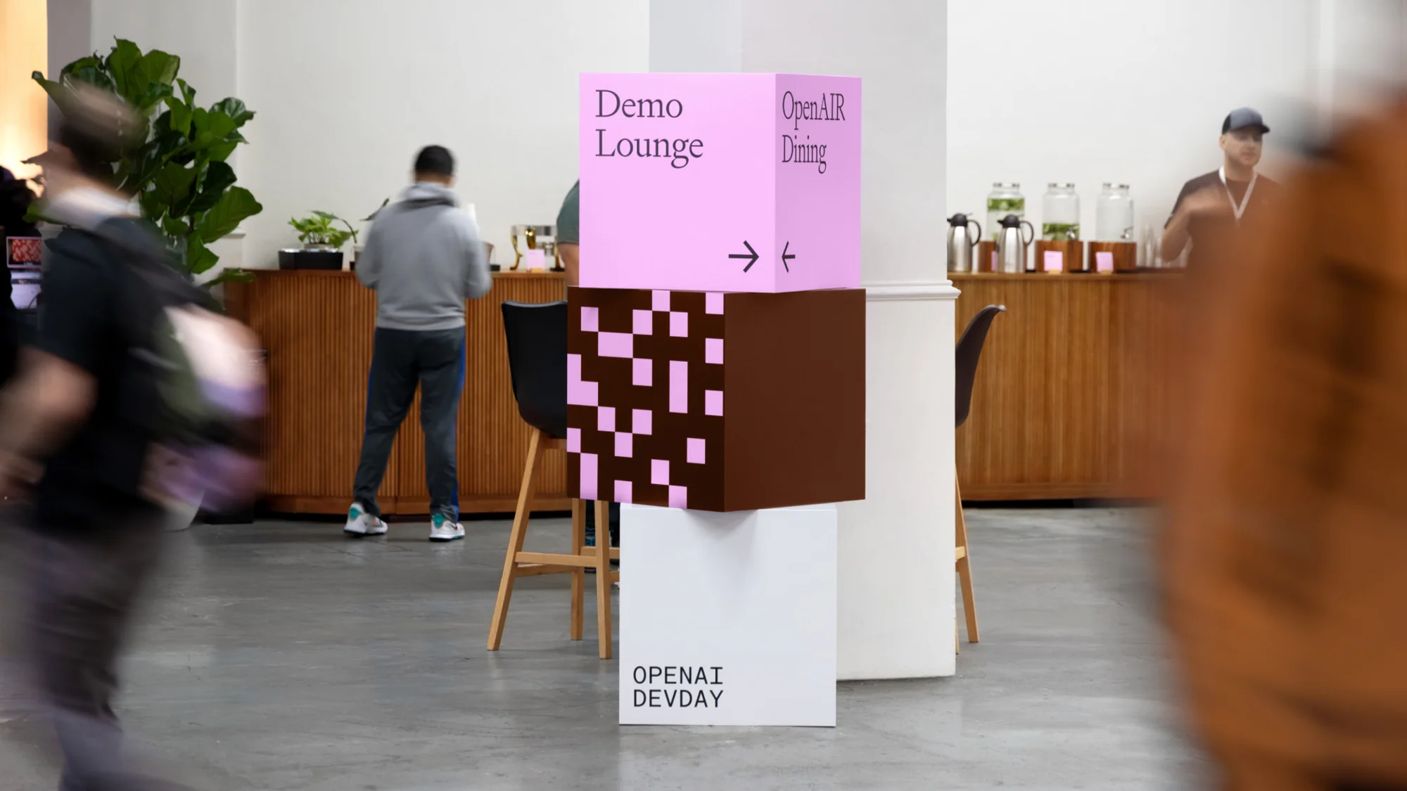 Event signage featuring a pink and brown cube stack, indicating directions for Demo Lounge and OpenAI Dining, inside a modern conference space.