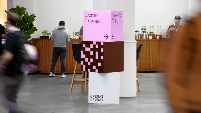 Event signage featuring a pink and brown cube stack, indicating directions for Demo Lounge and OpenAI Dining, inside a modern conference space.