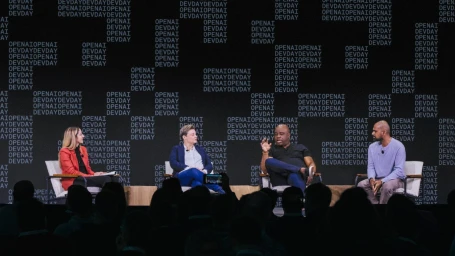 Four speakers seated on armchairs on a stage speaking to an audience in front of a backdrop reading OpenAI DevDay.