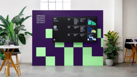 OpenAI DevDay event branding featuring a large purple and green pixelated backdrop with a digital directory screen