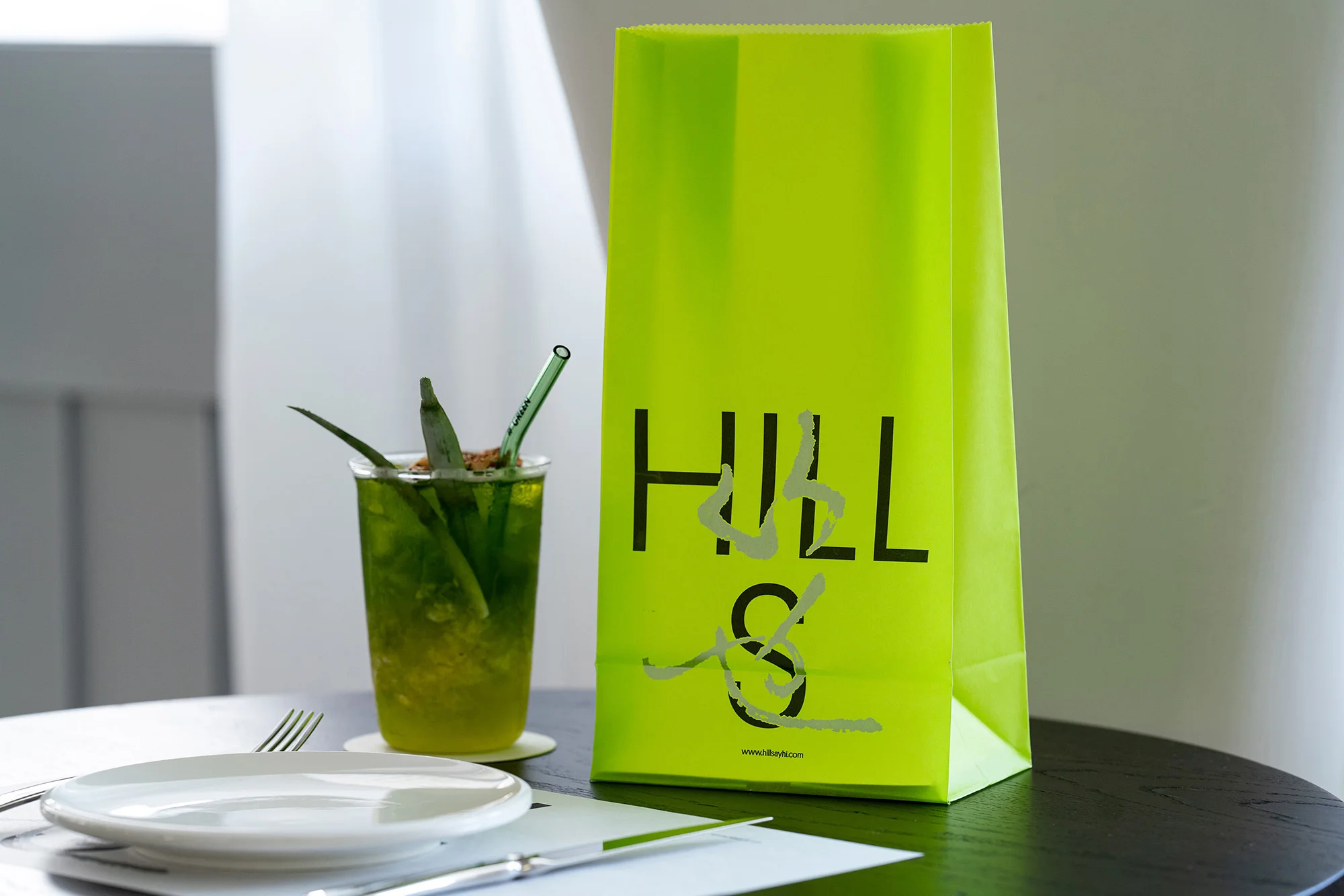 Beverage and bright green gift bag with logo sit on a black round table in a bright indoor setting with natural window light.