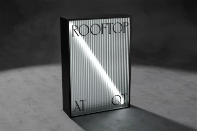 An illuminated rectangular lightbox sign with ribbed glass and serif typography spelling ROOFTOP AT QT, set against a dark grey concrete wall.