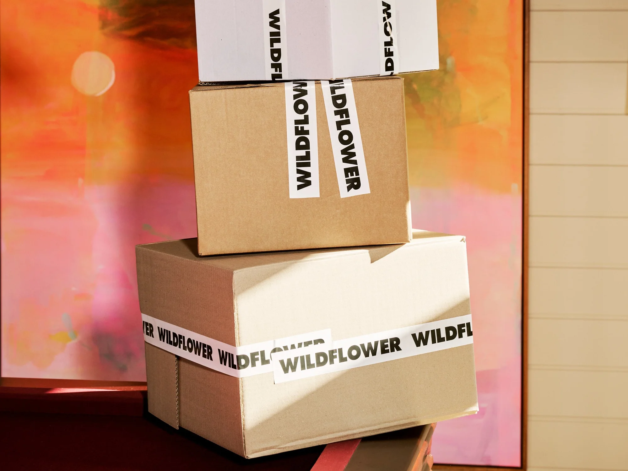 Stack of three cardboard shipping boxes labeled 'WILDFLOWER' on a wooden surface, set against a backdrop of abstract art and a paneled wall.