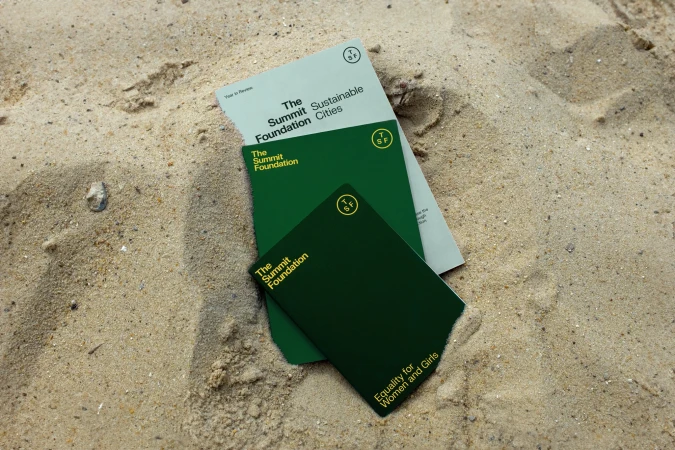 Three green and light-green colored books with The Summit Foundation logo displayed on a sandy beach in natural light.