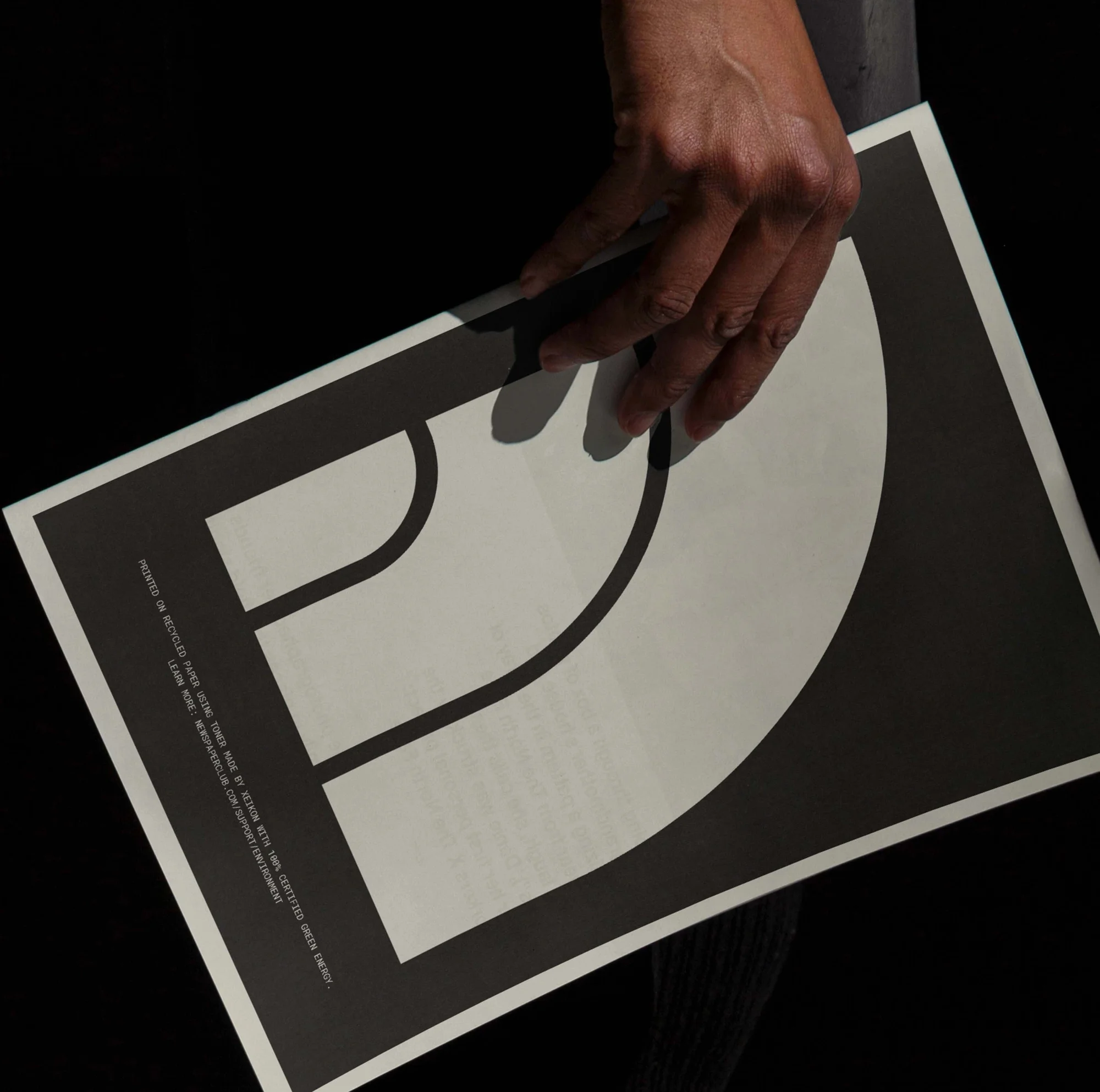 Adult's hand holds a white poster with a black graphic design, presented against a dark background with minimal visible details.
