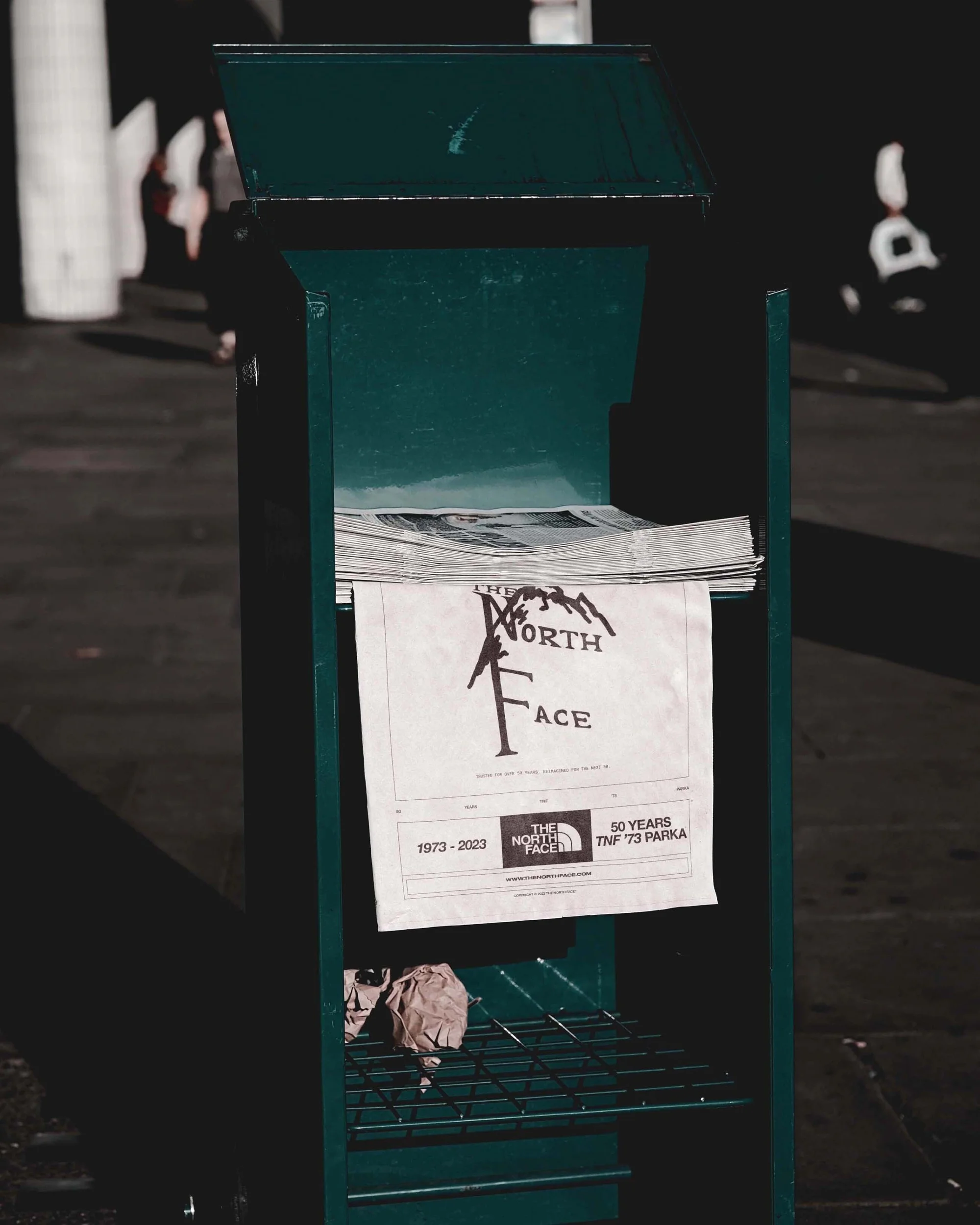 Close-up of a newspaper box in an outdoor urban setting, featuring a The North Face promotional flyer in a teal-colored metal box.