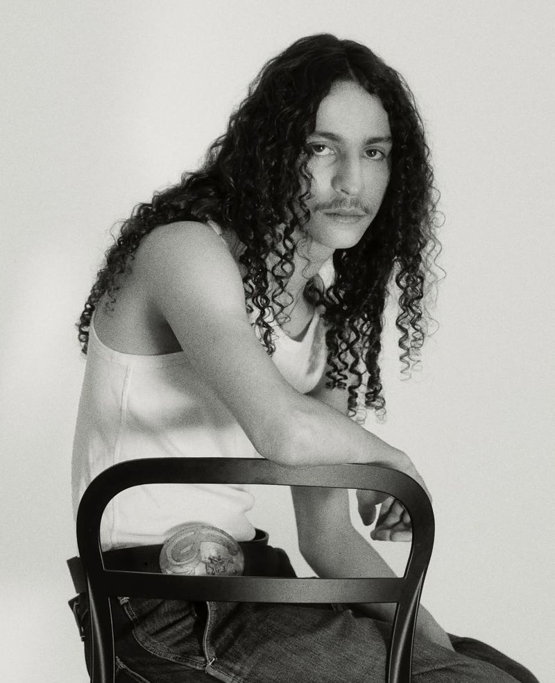 Black and white portrait of a person with long curly hair and a mustache sitting on a black chair in front of a plain white backdrop.