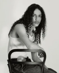 Monochrome portrait of Rayan Mazuel with long curly hair wearing a white tank top and dark trousers, seated behind a black chair.