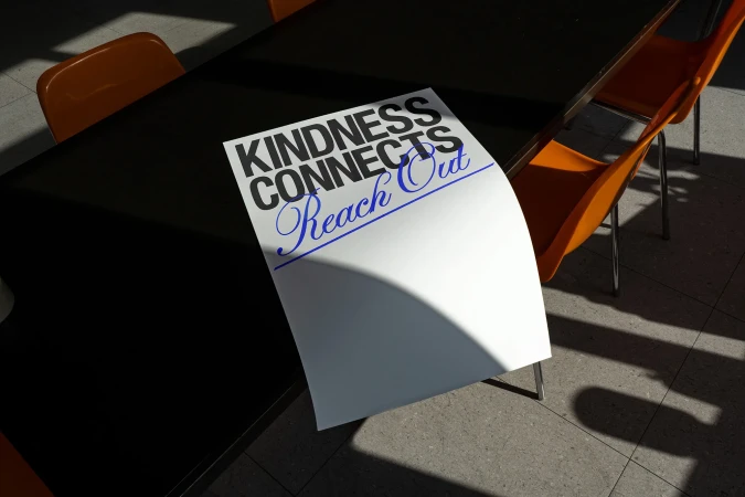 A sheet of paper with the text 'Kindness Connects Reach Out' is on a black table with orange chairs under natural lighting.