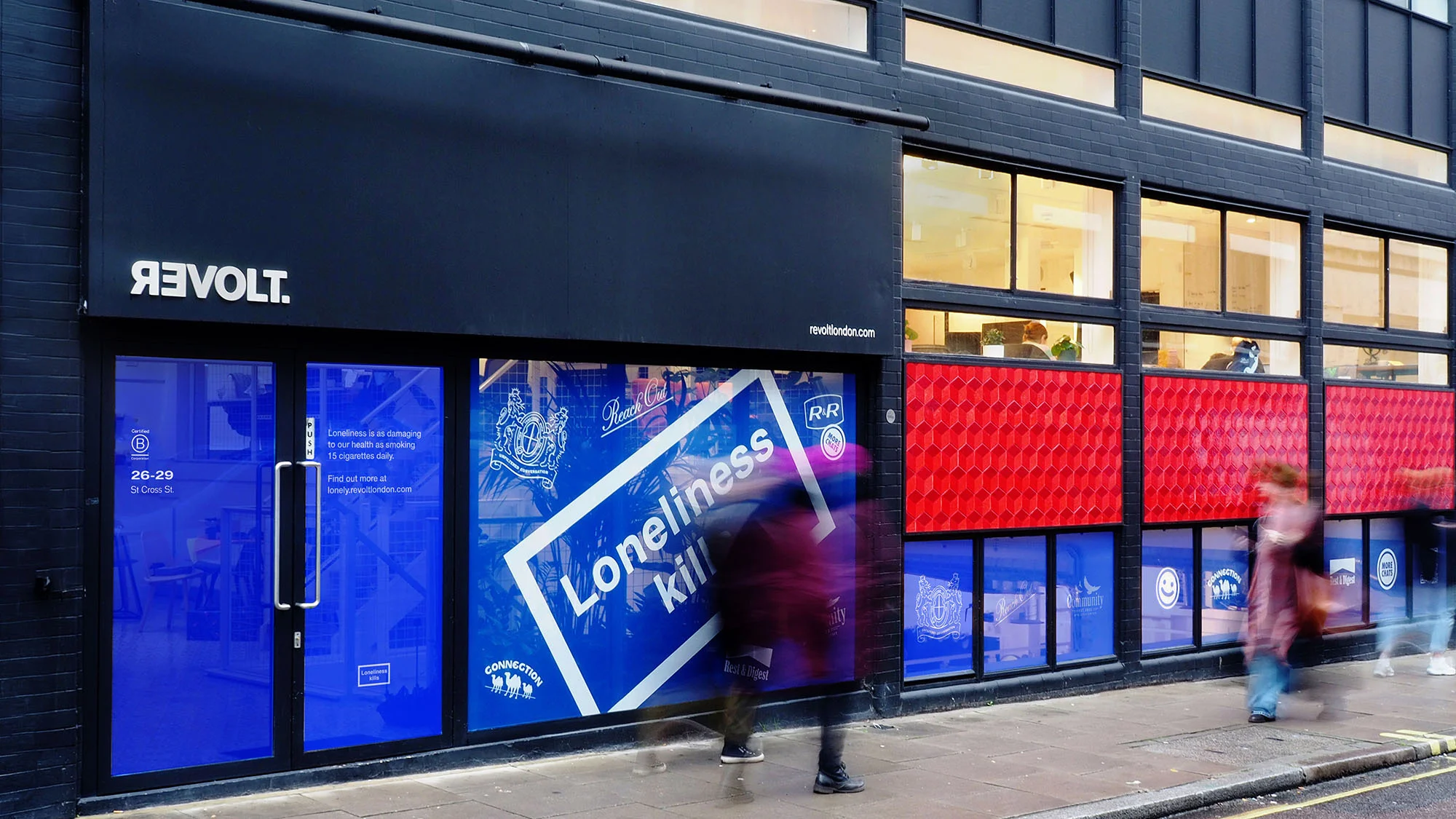 Exterior of REVOLT. building in London with a dark facade, blue-tinted windows, and graphic posters facing the street showing foot traffic.