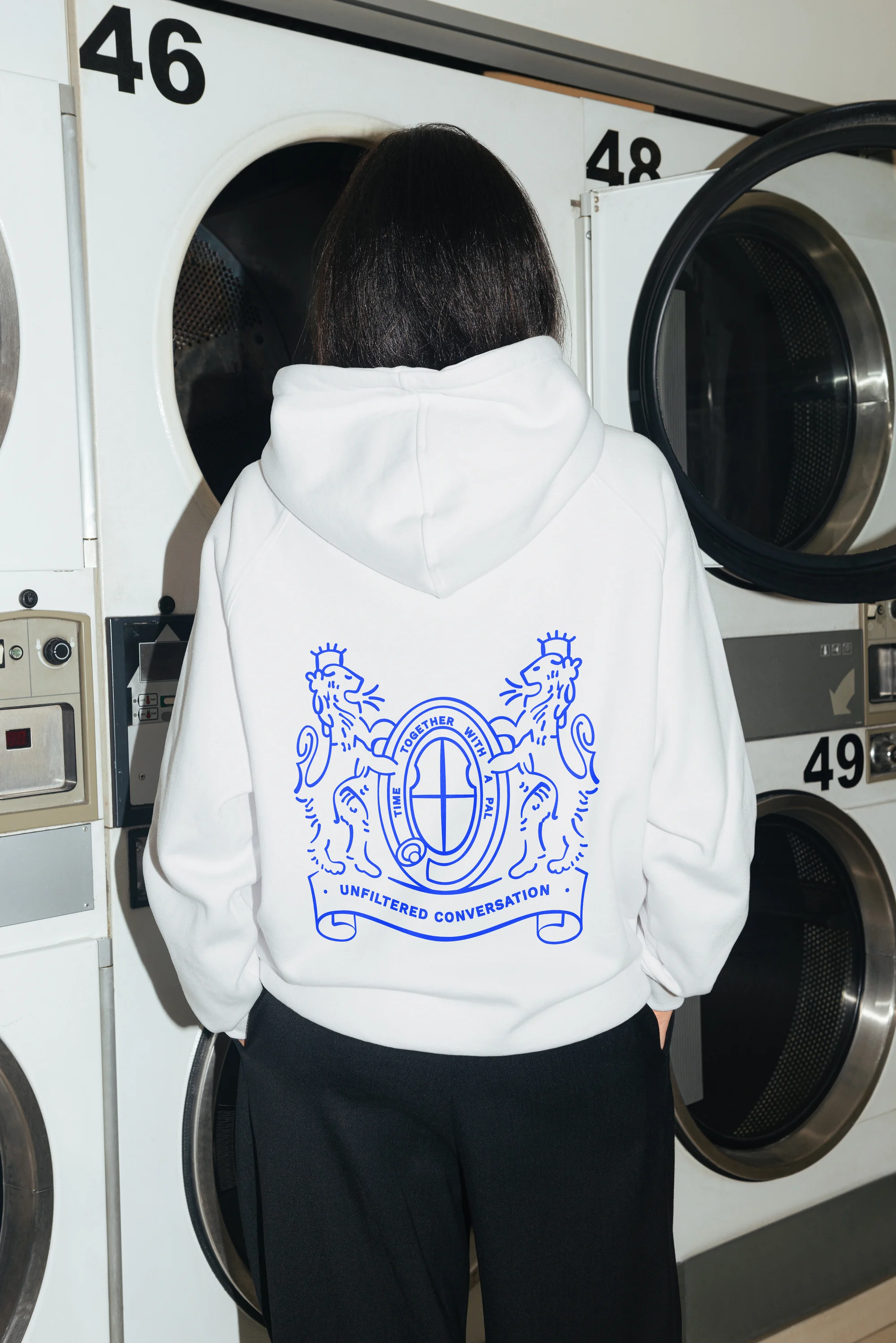 Woman in white hoodie with blue graphic stands in front of laundromat washing machines. Back view. Fashion, lifestyle photography.