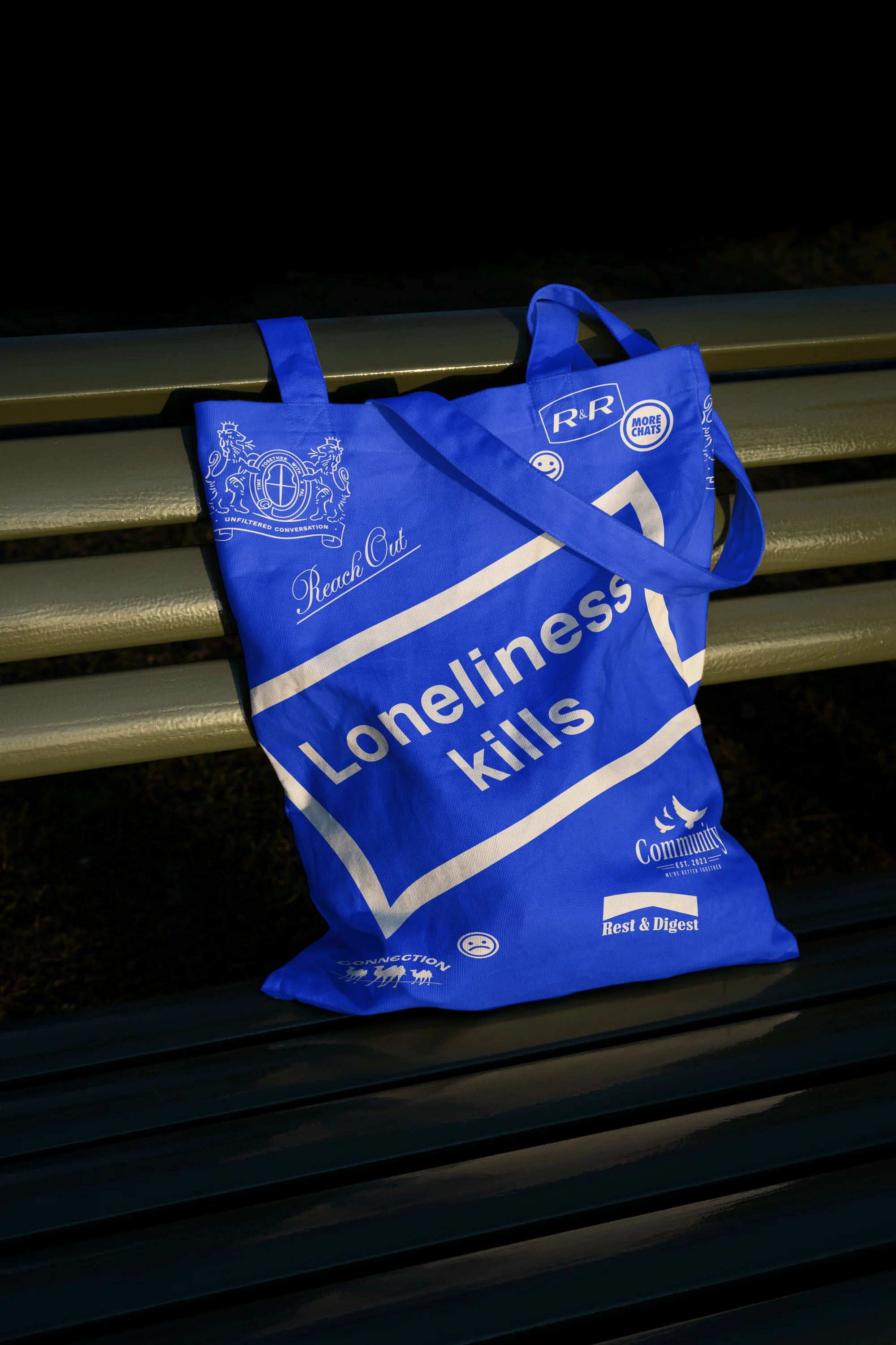Blue tote bag with white lettering 'Loneliness Kills' sitting on a green bench against a dark background in an outdoor setting.