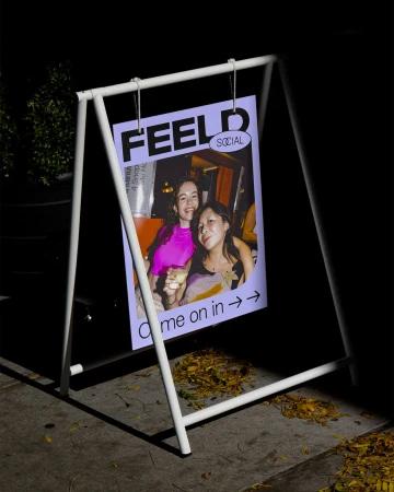 Sidewalk sign at night featuring a poster of two smiling young women inside a bar, inviting passersby to come on in.