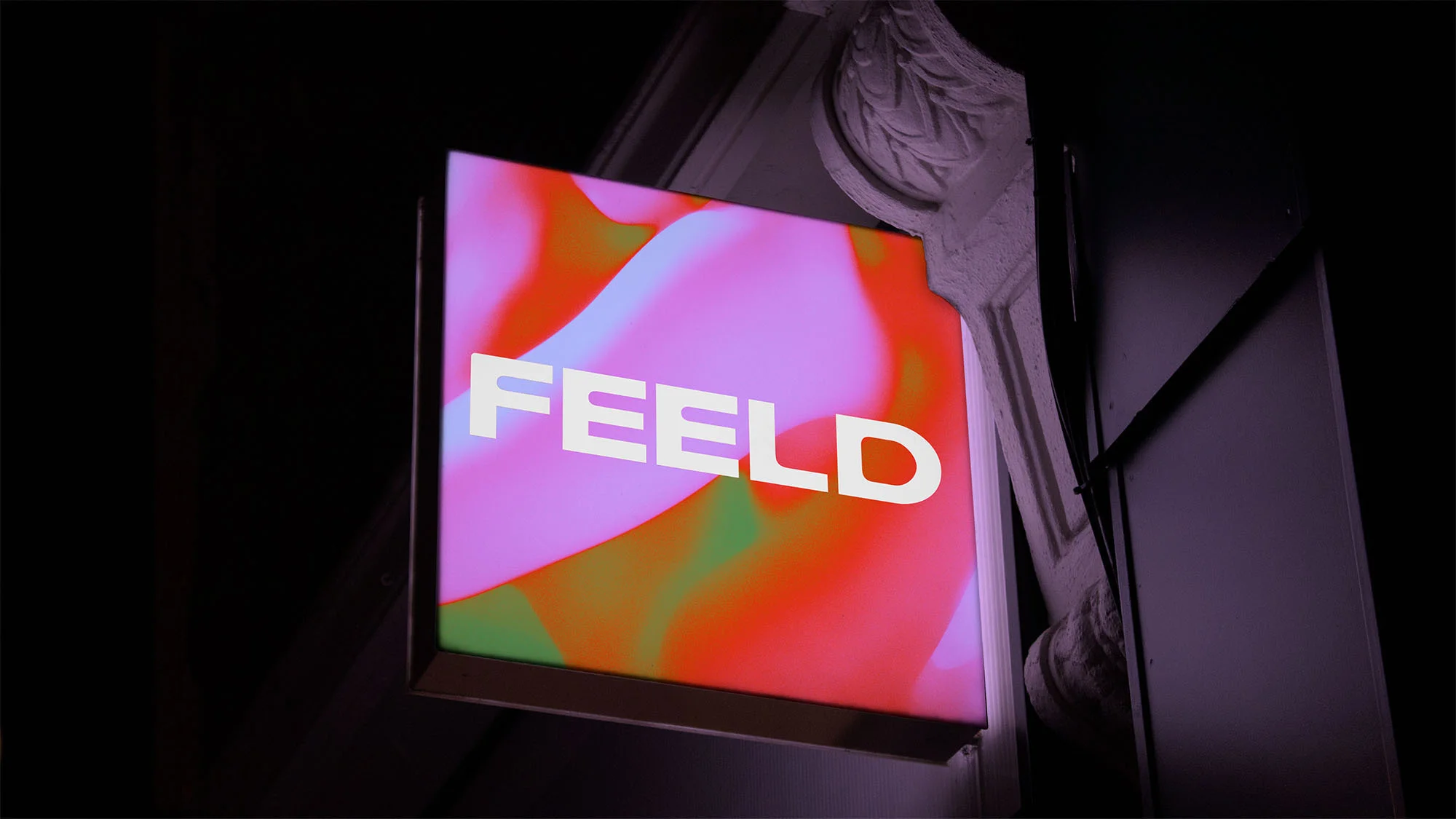 Illuminated business sign with the word "FEELD" in white letters against a pink, red, and green swirling abstract background.