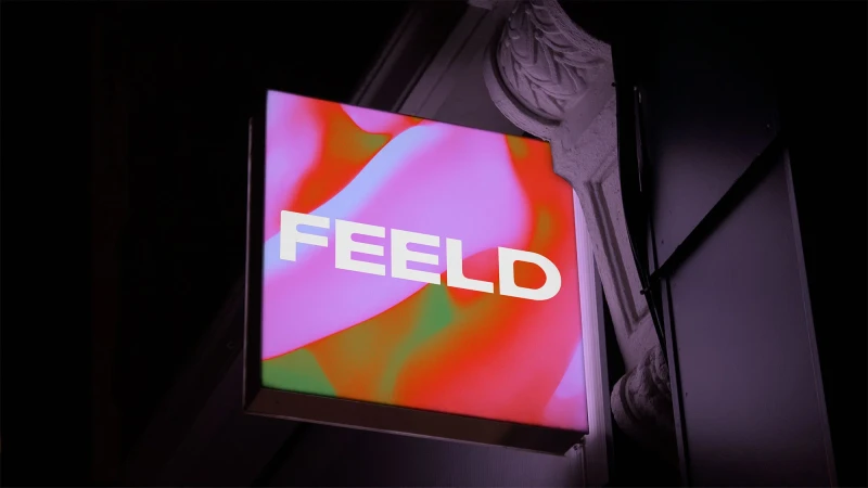 Illuminated business sign with the word "FEELD" in white letters against a pink, red, and green swirling abstract background.