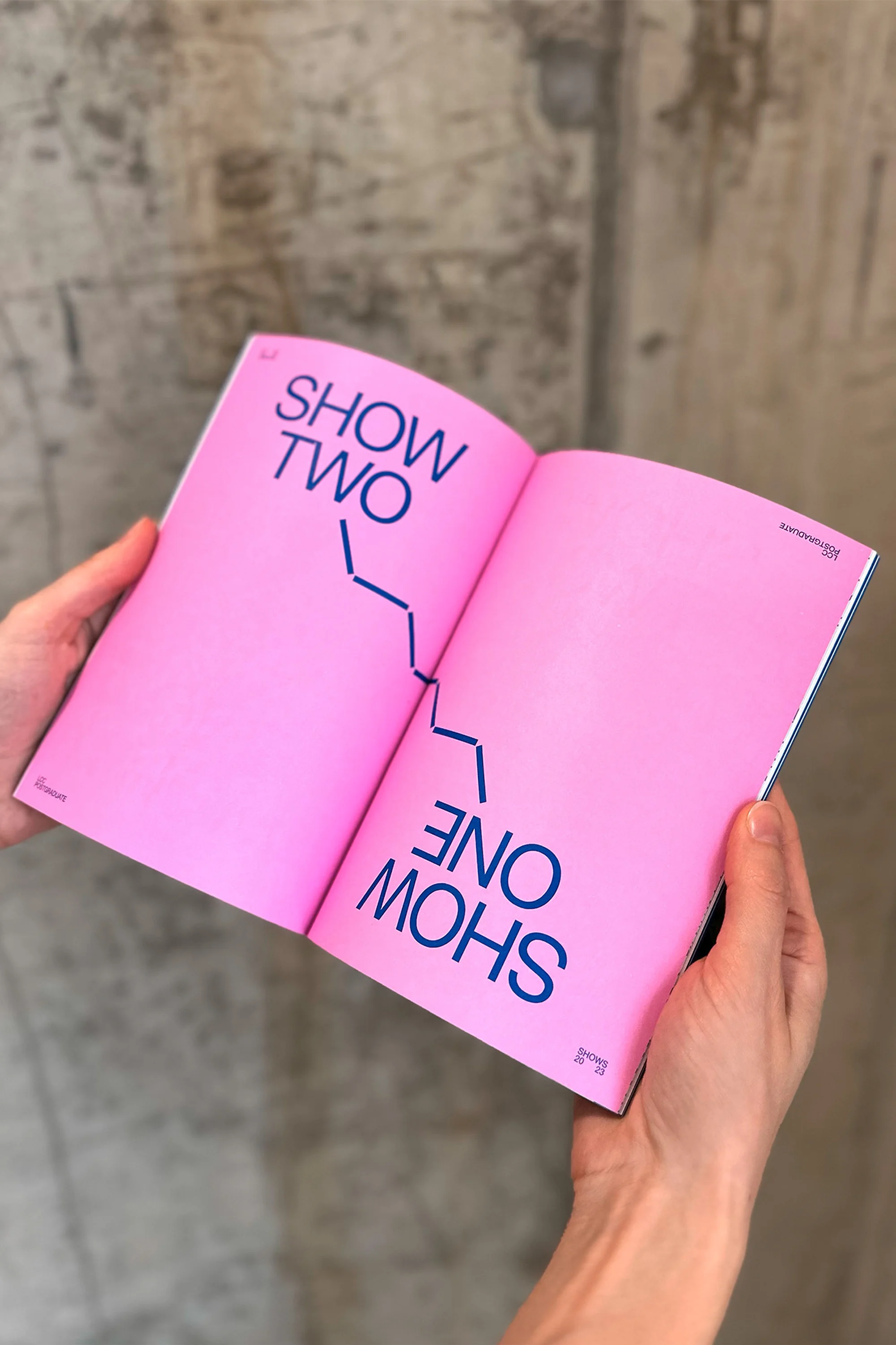 Hands hold open magazine with pink pages and blue text 'Show Two' and 'Show One' against a textured grey background, close-up.