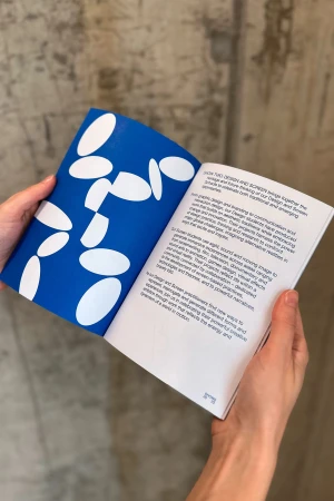 Hands hold open book with blue abstract design and white text on pages, showcasing graphic design and typography on a grey backdrop.
