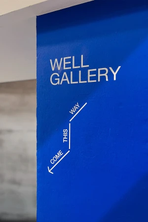Sign on a blue wall indicates direction to an art gallery with the words WELL GALLERY and an arrow graphic.