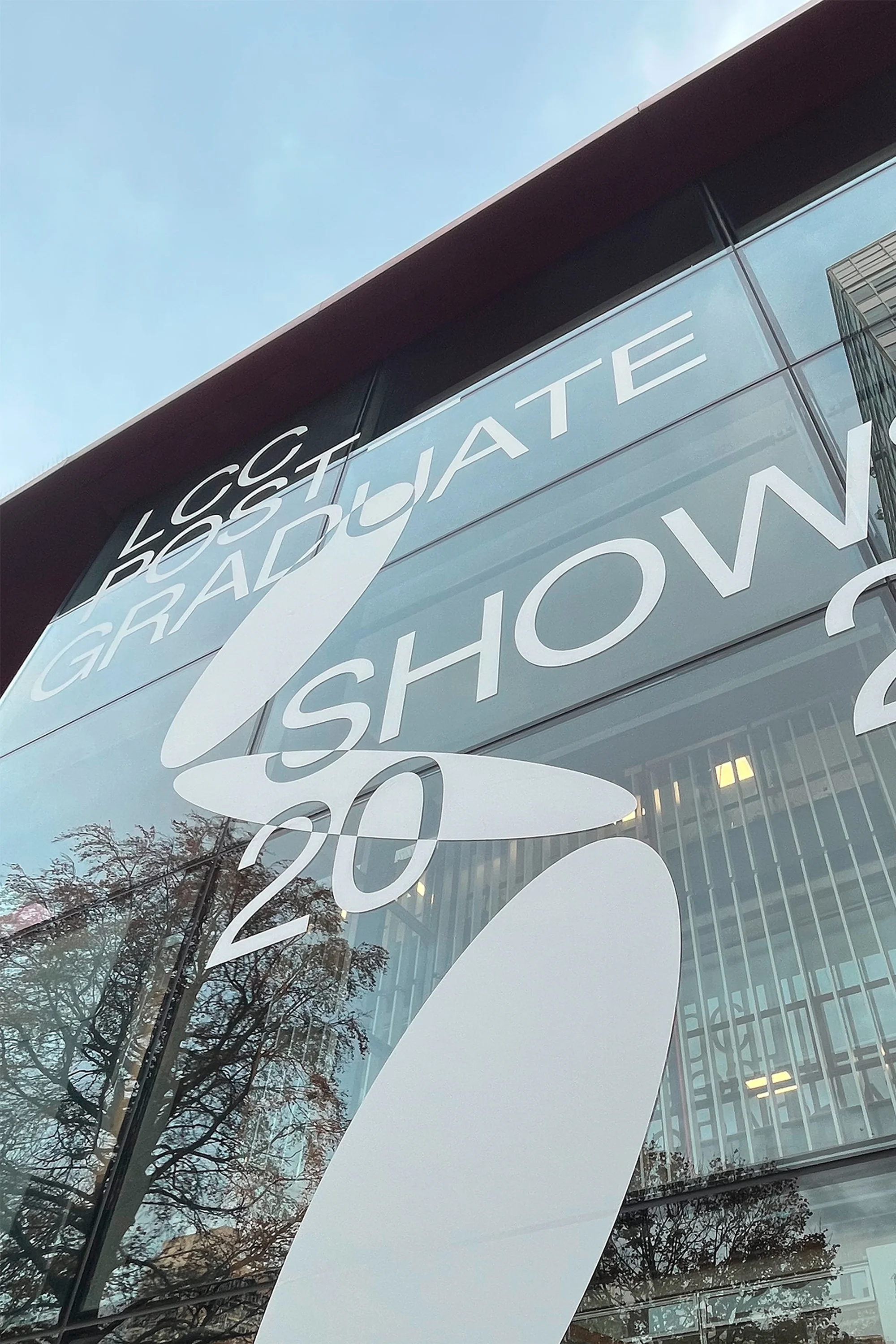 Building with the 'LCC Graduate Show 20' sign. Modern architecture with large glass windows reflecting trees and cloudy sky above.