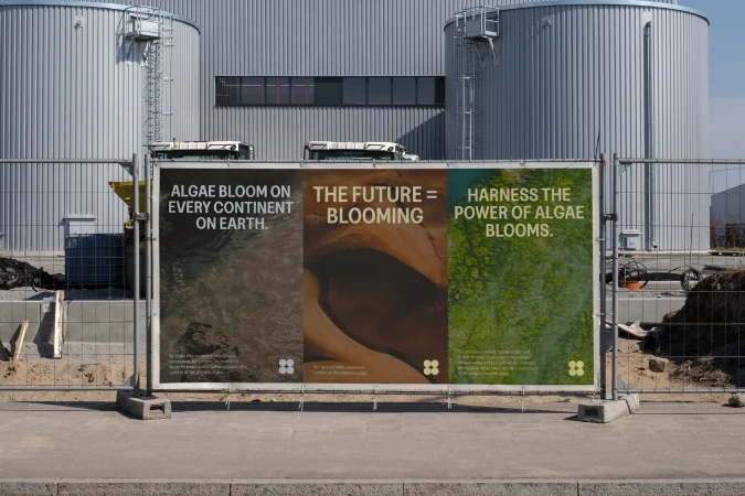 Outdoor view features three adjacent posters in metal frames displaying text and abstract algae imagery against an industrial background.