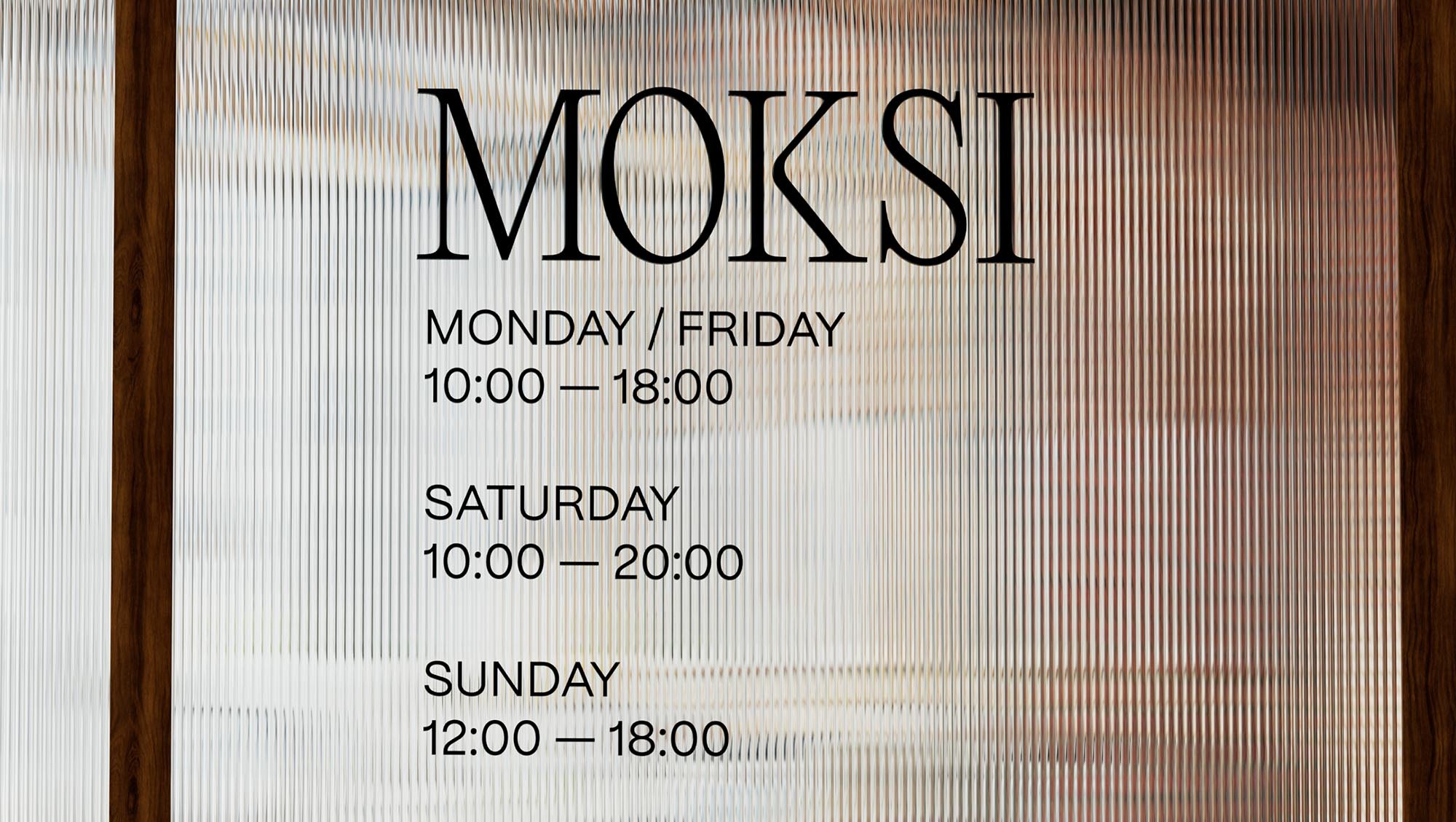 Store hours signage with name MOKSI, including Monday/Friday 10 AM–6 PM, Saturday 10 AM–8 PM, and Sunday 12 PM–6 PM against textured glass.