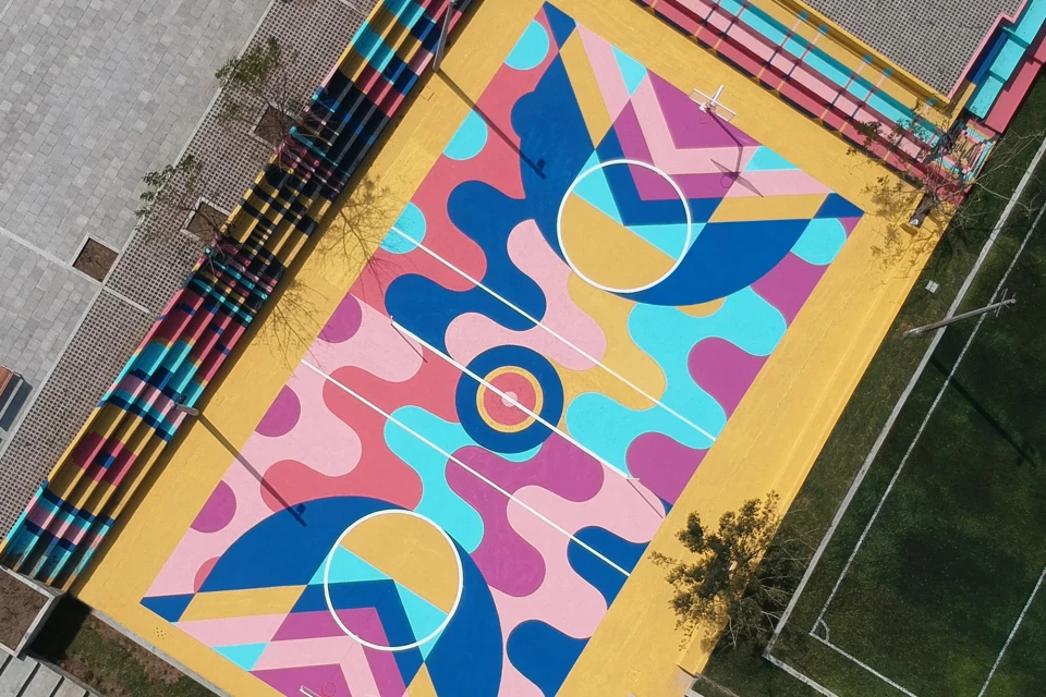 Aerial view of a colourful basketball court with abstract art patterns, surrounded by trees, grass, and a grey paved area.