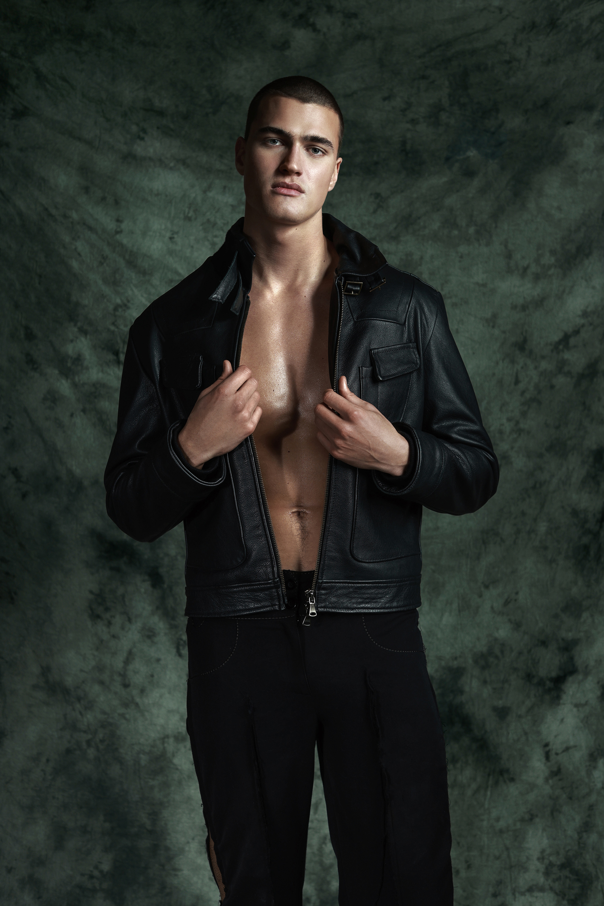 Portrait of a male fashion model with an open black leather jacket posing against a mottled green backdrop in a studio setting.
