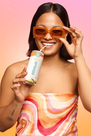 A woman in orange sunglasses smiles, holding a Nalu energy drink with a straw. She is wearing a multicolored scarf and the background is gradient orange to pink.