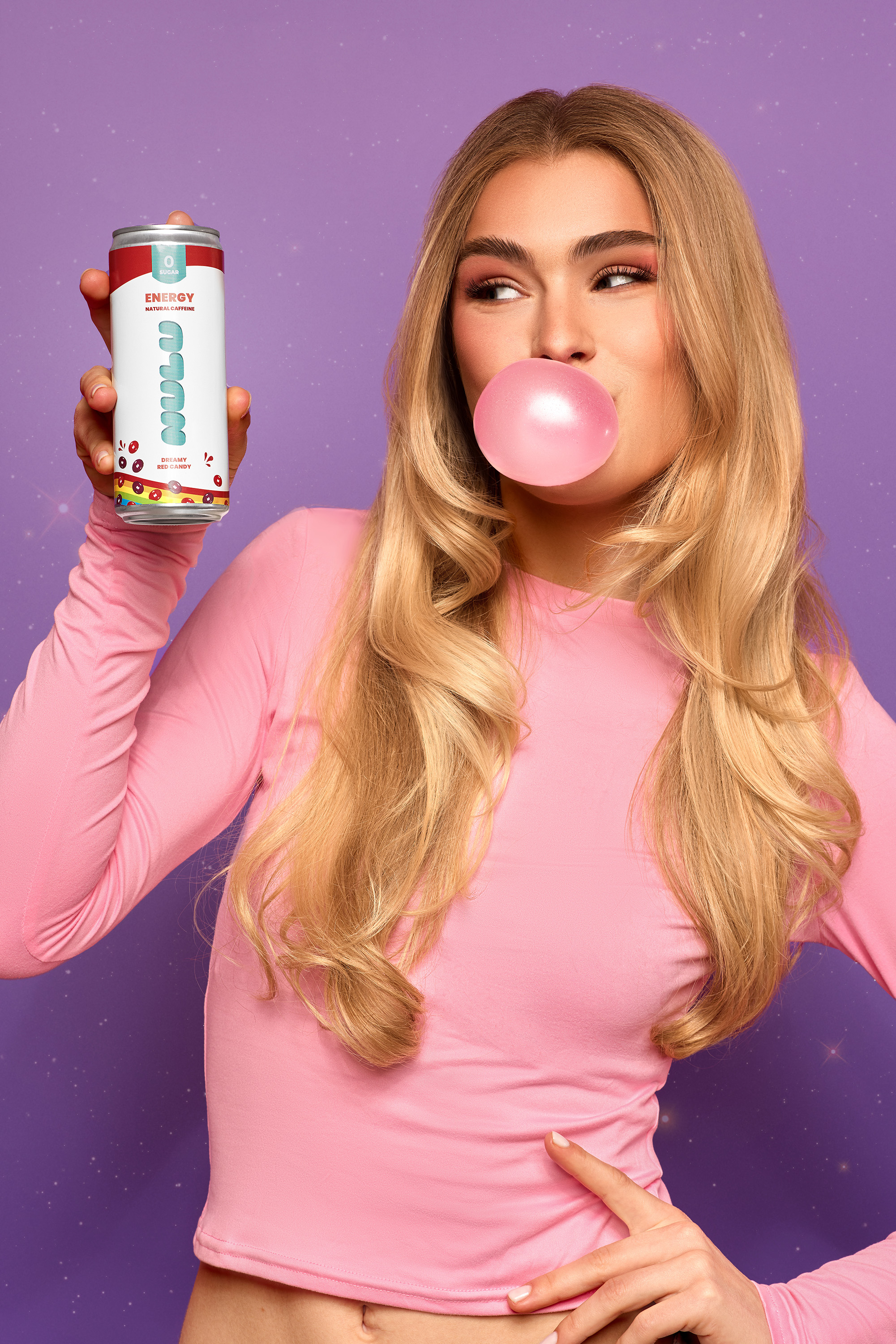 Woman blowing a pink bubblegum bubble holds an energy drink against a purple background. The drink is white with colorful fruit graphics.