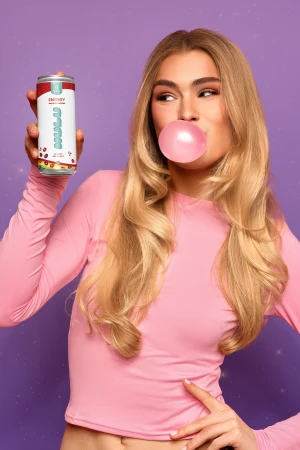 Woman blowing a pink bubblegum bubble holds an energy drink against a purple background. The drink is white with colorful fruit graphics.