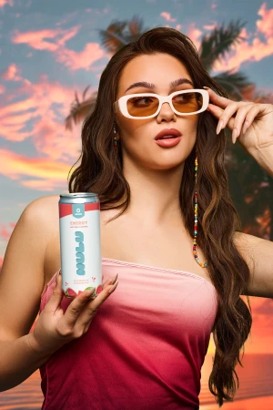 Attractive woman in sunglasses holds a Nulu energy drink against a backdrop of palm trees and a vibrant sunset sky.