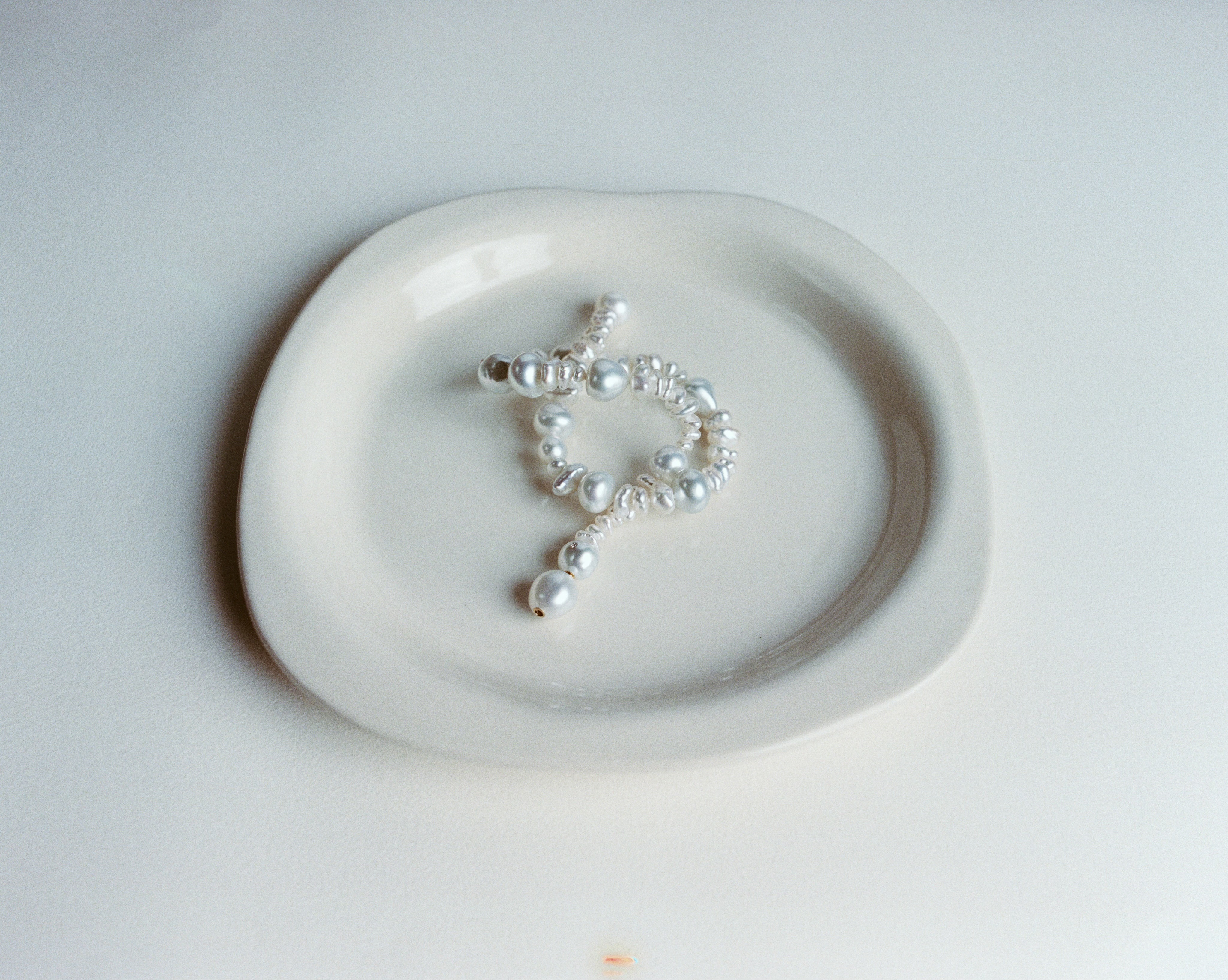 A strand of freshwater pearls is arranged on a white ceramic jewelry plate, photographed in a bright studio setting.