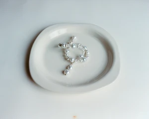 An intricate pearl necklace arranged in a loop on a white ceramic dish against a neutral background