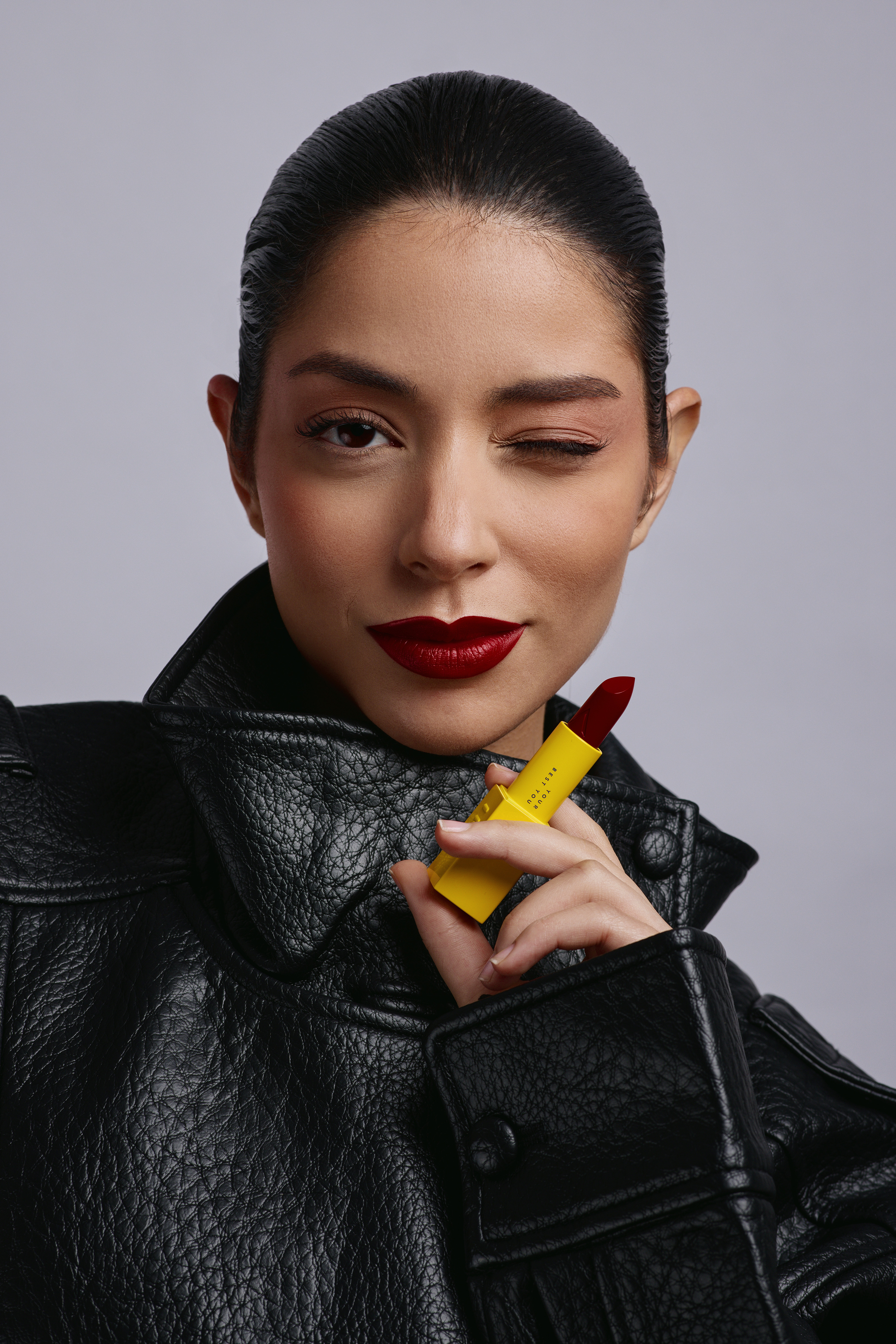 Attractive brunette adult woman wearing a black leather jacket winking at the camera and holding a yellow lipstick tube with red lipstick.