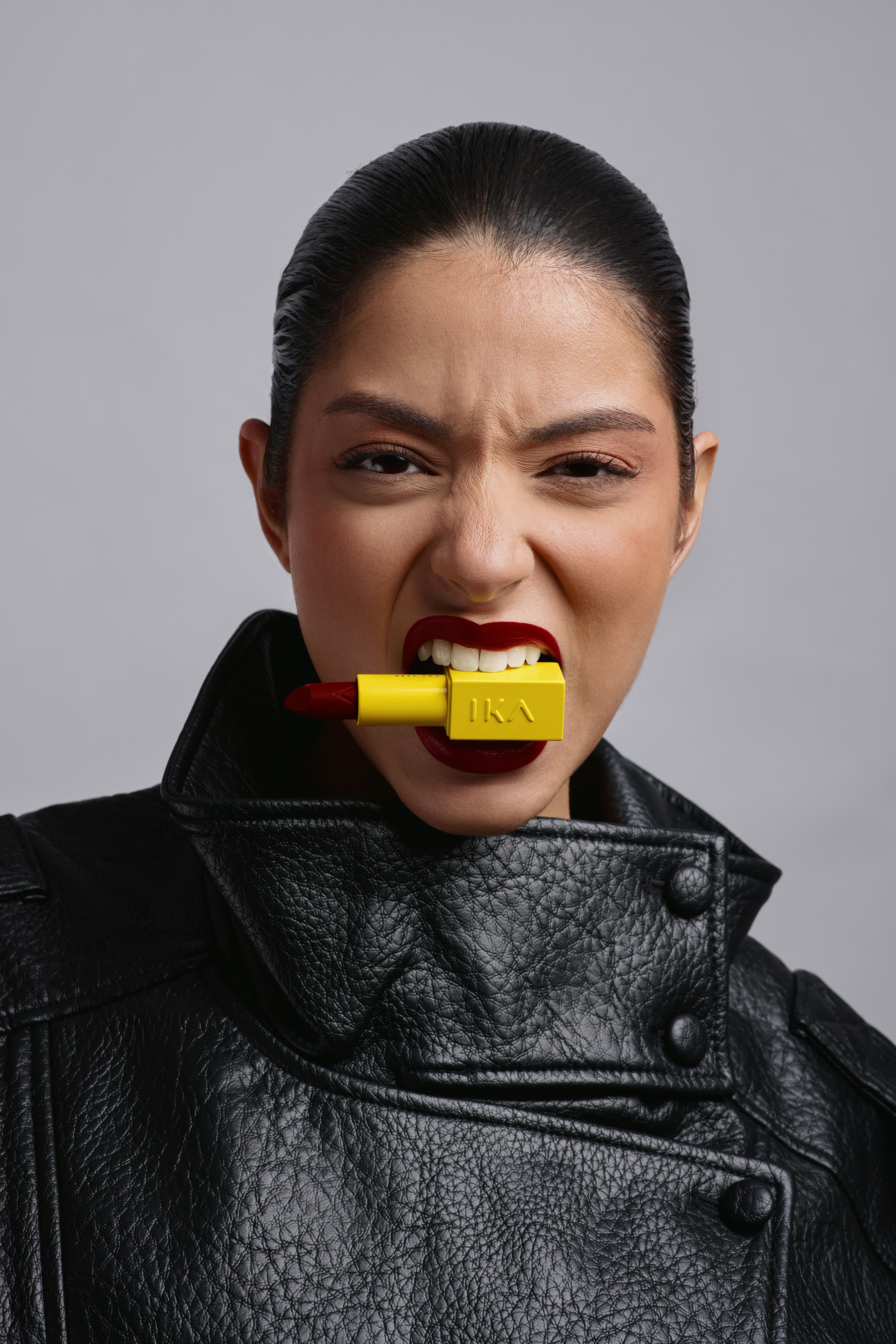 Close-up of a fierce woman in a leather jacket biting on a yellow lipstick. Bold beauty statement in a studio setting, featuring makeup.