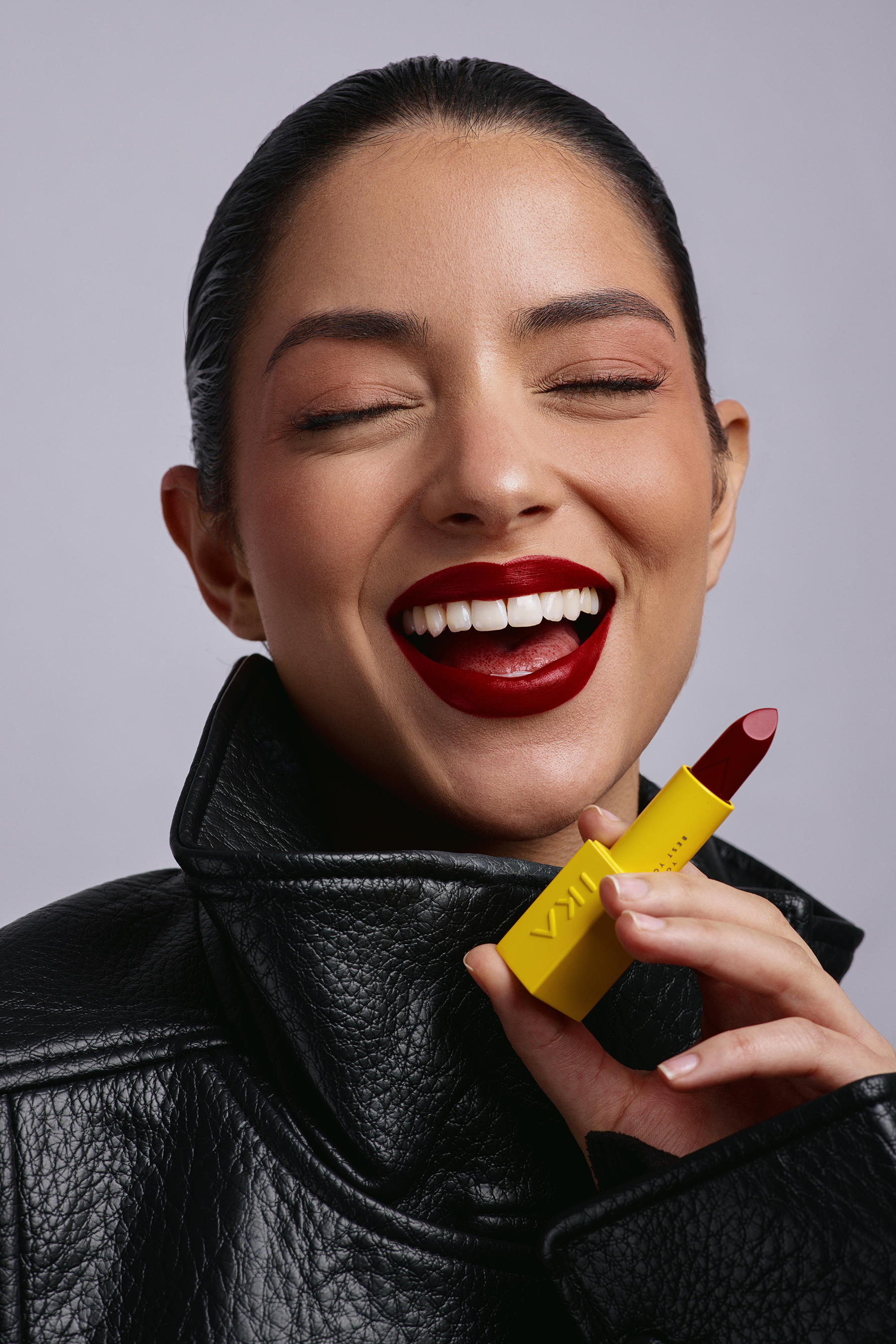 Close-up of a smiling woman with dark red lipstick, holding a yellow lipstick tube with visible brand, against a light gray background.