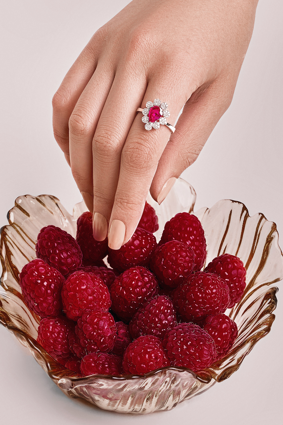 Close-up of a hand adorned with a ruby and diamond ring reaching into a bowl of fresh raspberries against a light background.