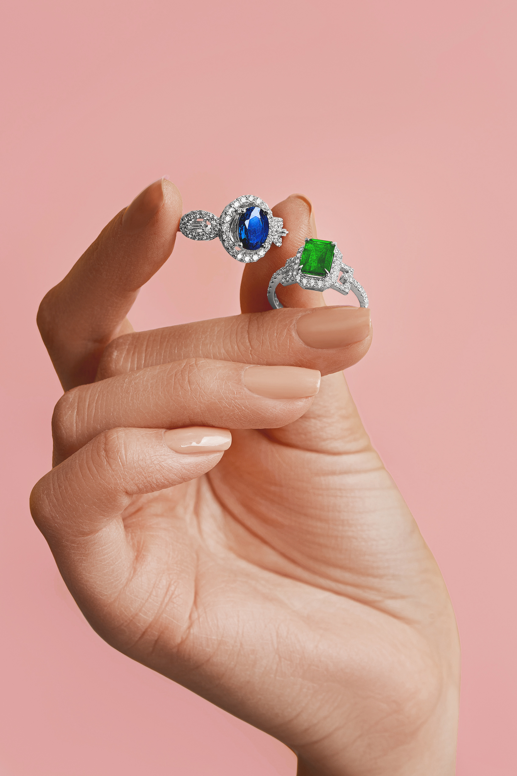 Close-up of a hand holding two gemstone rings: one with a blue stone, the other with a green stone, set against a pink background.
