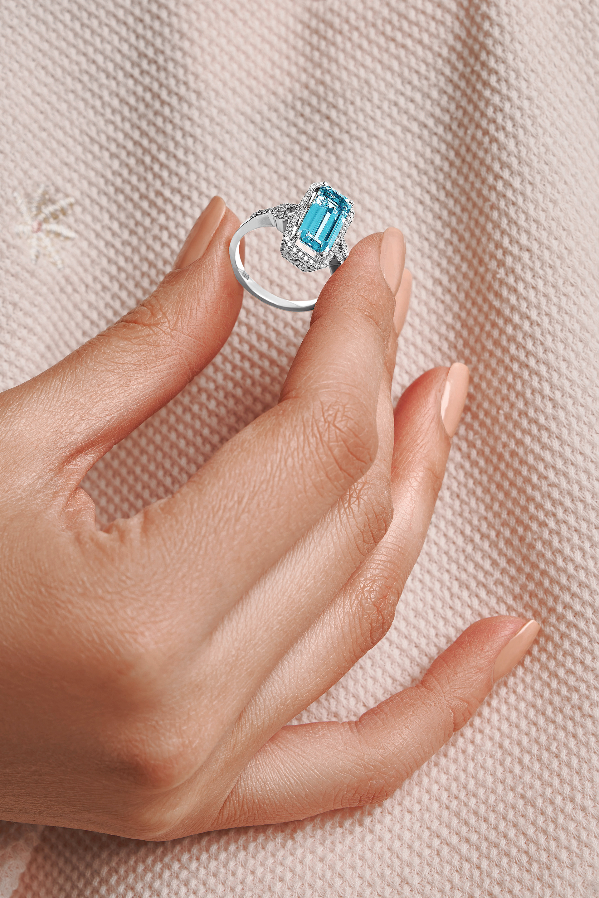 Close-up of a woman's manicured hand holding a sparkling silver ring featuring a rectangular aquamarine gemstone and diamond halo.