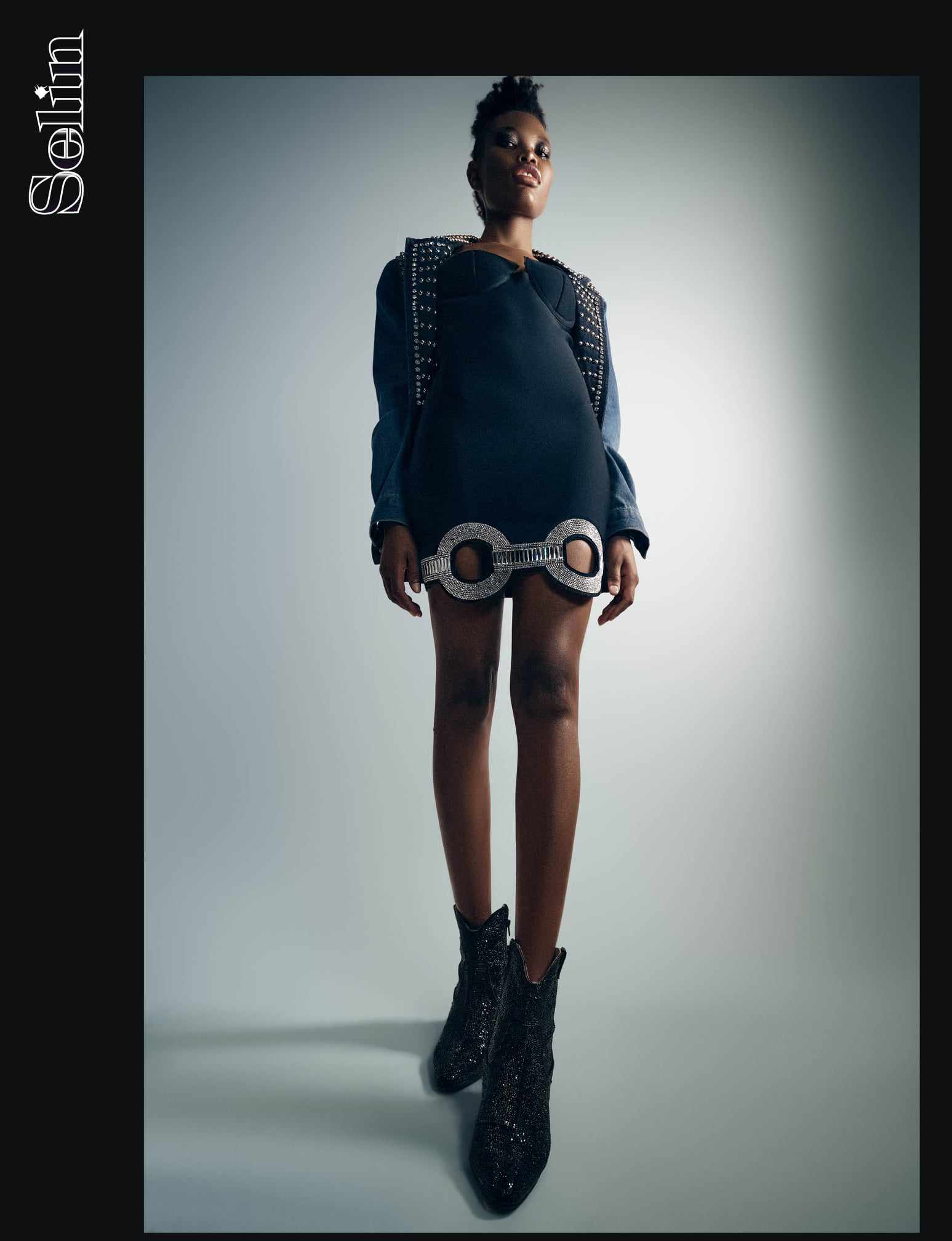 Low angle full-length shot of a Black woman wearing a black mini dress and embellished jacket against a gradient background.
