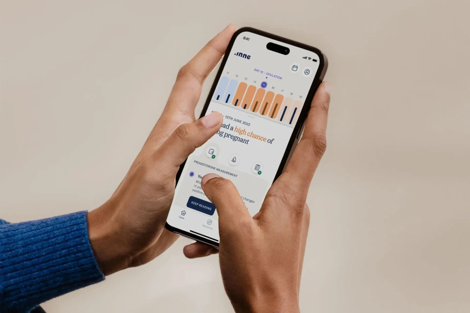 Close-up of a woman's hands holding a smartphone displaying a fertility app, showcasing graphs and data on a light beige background.