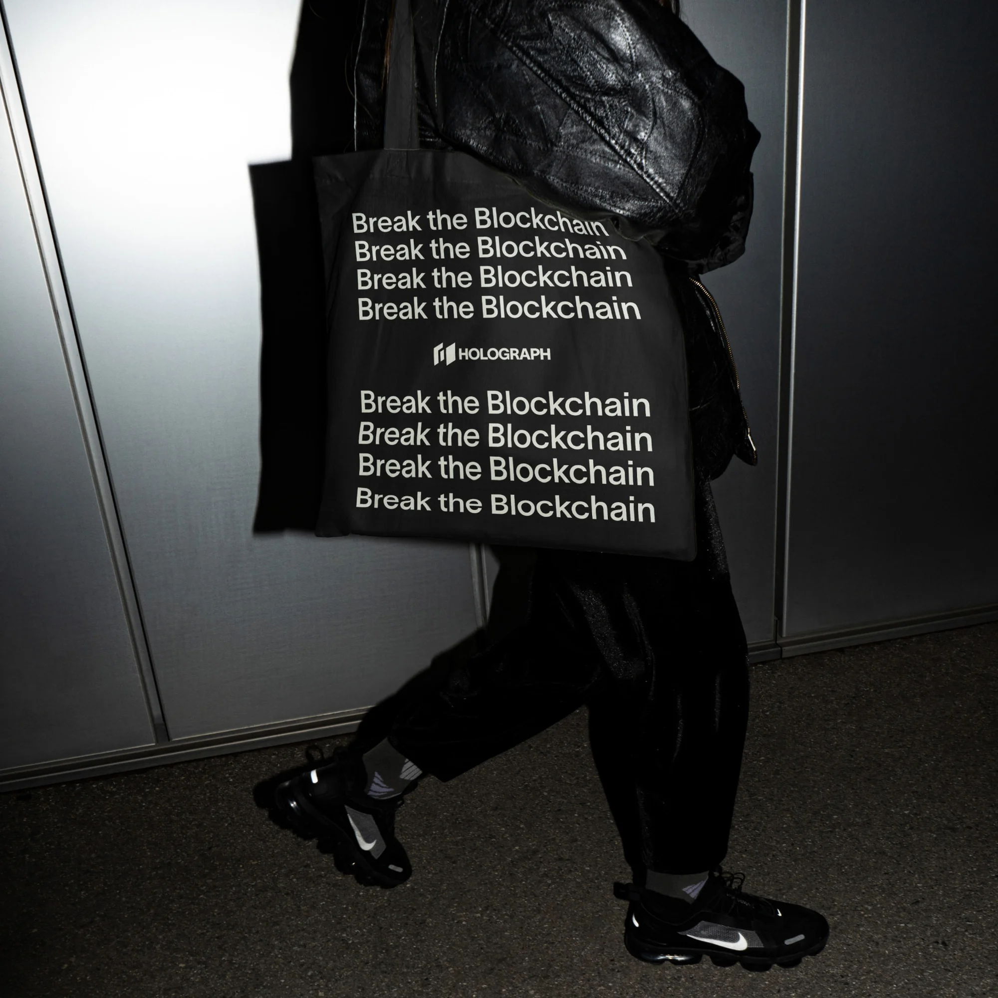 A person is walking wearing black sneakers with a white logo and carrying a black tote bag that says "Break the Blockchain."