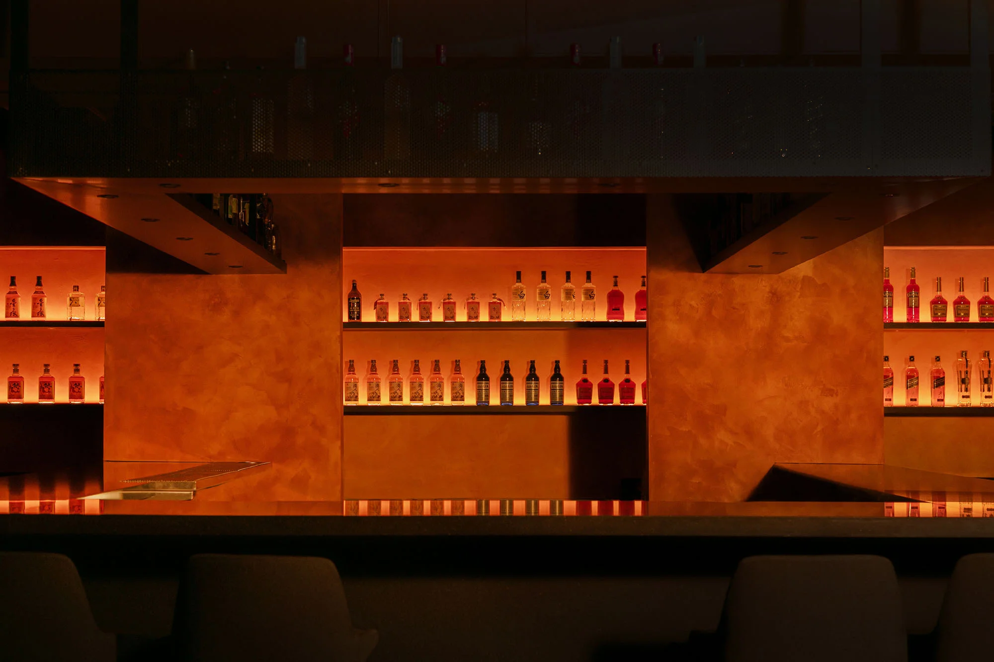 Illuminated bar interior with liquor bottles displayed on shelves. Bar counter and stools in the foreground, warm orange lighting.