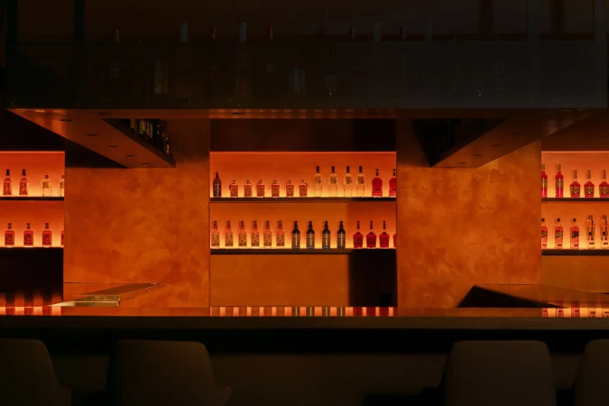 Illuminated bar interior with liquor bottles displayed on shelves. Bar counter and stools in the foreground, warm orange lighting.