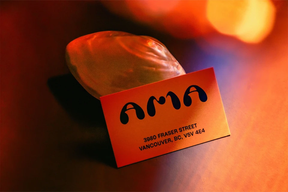 Business card with 'AMA' and address in Vancouver, BC, beside a small shell. Warm lighting creates an inviting ambiance.