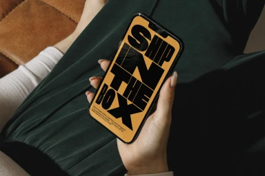 Close-up of hands holding a smartphone displaying a yellow screen with bold black typography reading Ship in the Box against dark green clothing