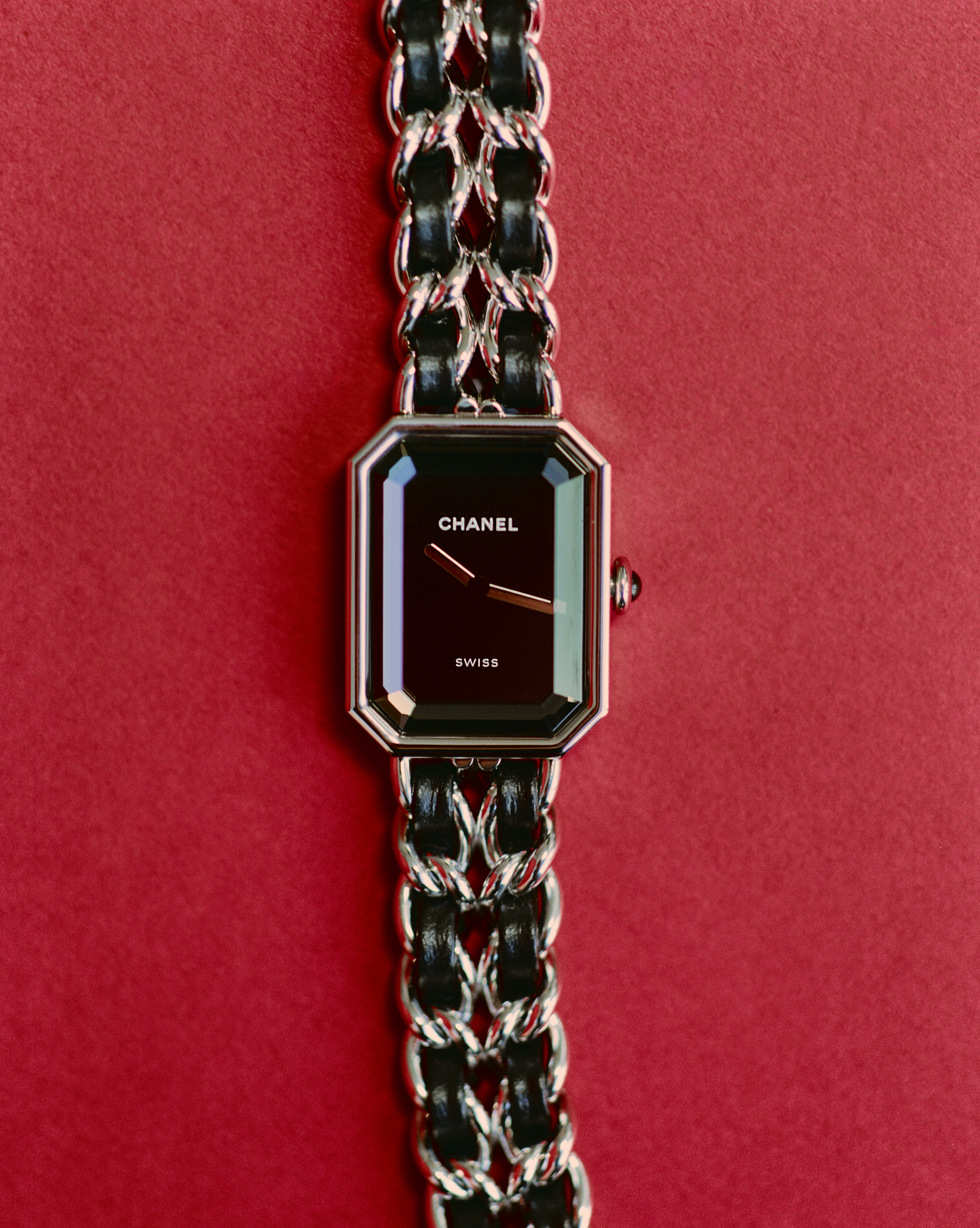 Chanel Première watch with black dial and interwoven leather and metal chain band on a textured red background.
