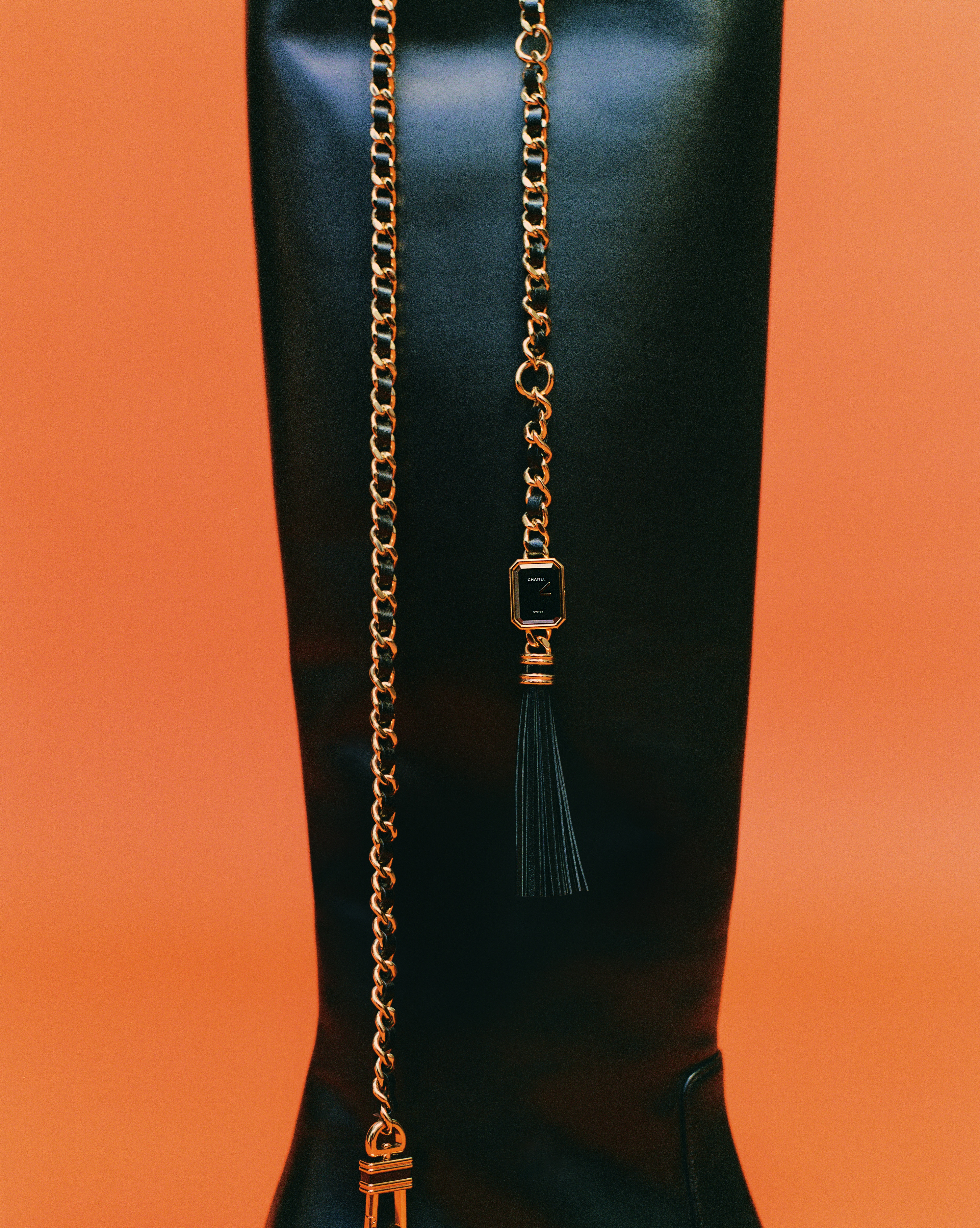 Black leather boot with dangling gold chain and black tassel on an orange backdrop, showcasing fashion accessory details and texture.