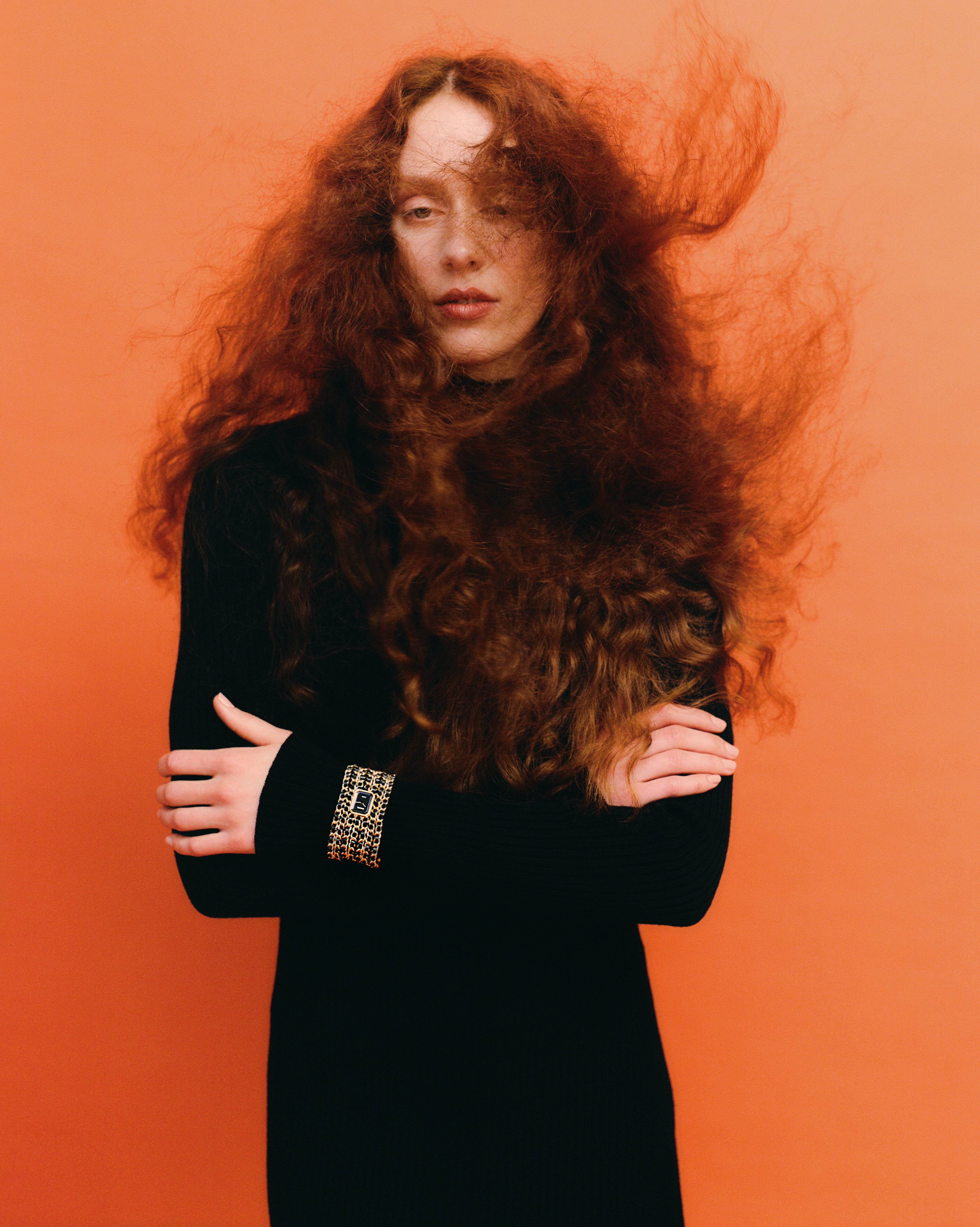 Portrait of a red-haired adult female wearing a black dress against an orange backdrop, creating a striking fashion or beauty image.